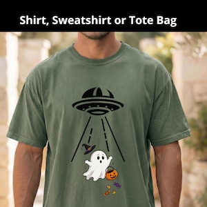 May include: An olive green short-sleeved t-shirt featuring a black graphic of a UFO beaming down a white ghost wearing a purple witch hat. The ghost is holding an orange jack-o'-lantern bucket, with small colorful candies scattered below.