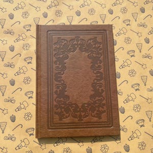 Puede incluir: Un diario encuadernado en cuero marrón con un marco decorativo en relieve en la portada. El diario es rectangular y cerrado, con una superficie texturizada. El fondo es un papel de regalo beige con un patrón repetido de ilustraciones navideñas.