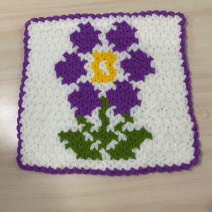 Könnte beinhalten: Ein quadratischer, handgefertigter Häkelartikel mit einem großen Blumendesign. Die Blume hat lila Blütenblätter, eine gelbe Mitte und grüne Blätter. Der Hintergrund ist weiß und der Rand ist lila.