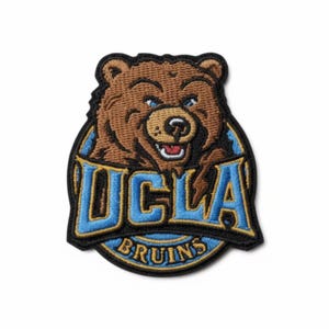 Puede incluir: Parche bordado con el logotipo de los Bruins de la UCLA. Presenta un oso pardo con ojos azules y una expresión sonriente. El texto "UCLA" está en azul y dorado, y "BRUINS" está en azul.