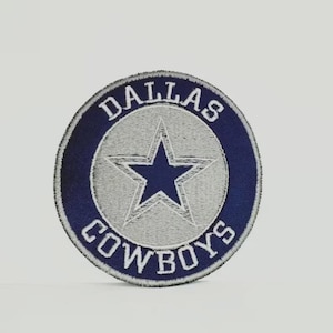May include: Embroidered patch featuring the Dallas Cowboys logo. The patch is circular, with a navy blue border and a gray center. The words "DALLAS COWBOYS" are embroidered around the edge. A navy blue star is in the center.