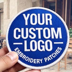Puede incluir: Parche bordado redondo azul con texto blanco "YOUR CUSTOM LOGO EMBROIDERY PATCHES". El parche tiene un borde blanco y está sostenido por una mano.