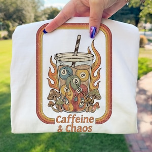 May include: White sweatshirt featuring a retro graphic of a coffee drink with billiard balls, flames, and mushrooms. The text "Caffeine & Chaos" is printed below the graphic. The design is framed in a retro-style border.
