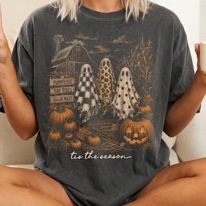 May include: A charcoal gray t-shirt featuring a graphic print of three ghosts in front of a barn, pumpkins, and corn stalks. The text reads PUMPKINS, HAY RIDES, CORN MAZE, and tis the season. The ghosts have checkered, leopard, and polka dot patterns.