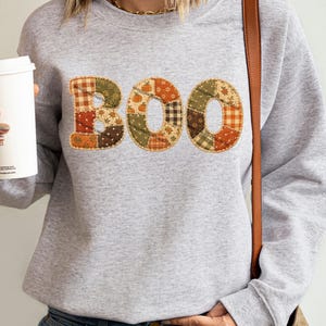 May include: A light gray crewneck sweatshirt featuring the word BOO printed in a patchwork style. The letters are filled with various autumn-themed patterns including orange pumpkins, brown plaid, and green floral motifs on a textured background.