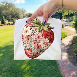 Può includere: Felpa bianca con un grande design a fragola ricamata. La fragola è composta da tessuto patchwork nei toni del rosso, rosa e verde, con dettagli floreali. Il testo "Passy Peach Mercantile" è stampato sulla felpa.