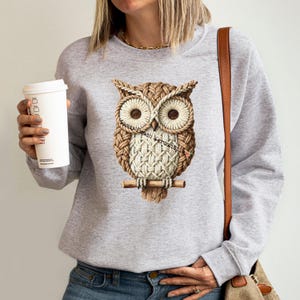 May include: A light gray sweatshirt featuring a detailed owl design. The owl is rendered in shades of brown and cream, with a textured appearance, perched on a wooden branch. The text "Sassy Peach Mercantile" is printed on the owl. The person is holding a white coffee cup.