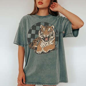 Puede incluir: Camiseta verde descolorida con un gr&aacute;fico de tigre. El tigre es marr&oacute;n y blanco, sobre un c&iacute;rculo a cuadros negro y gris. La camiseta tiene mangas cortas y un corte holgado.