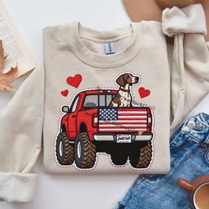 May include: A beige sweatshirt featuring an embroidered design of a red truck with a dog in the bed, an American flag, and red hearts. The truck has large tires and a license plate that says "patriot".