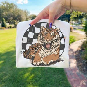Puede incluir: Camiseta blanca con un gr&aacute;fico de tigre rugiente en tonos marr&oacute;n y beige, sobre un c&iacute;rculo a cuadros blanco y negro. La palabra "Savage" est&aacute; impresa en la camiseta.