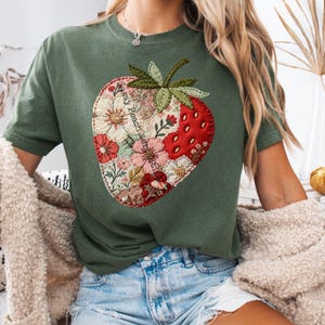 Könnte beinhalten: Grünes T-Shirt mit einem großen Erdbeer-Design. Die Erdbeere besteht aus Stoffflicken mit floralen Mustern und roten Abschnitten. Der Text "Sassy Peachwere" ist auf die Erdbeere gedruckt. Das Shirt hat kurze Ärmel.