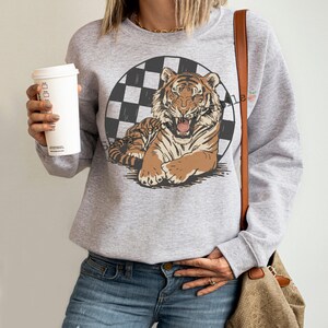 Puede incluir: Una sudadera gris claro con un gr&aacute;fico de tigre. El tigre es marr&oacute;n y beige, sobre un c&iacute;rculo a cuadros blanco y negro. La sudadera tiene mangas largas y cuello redondo.