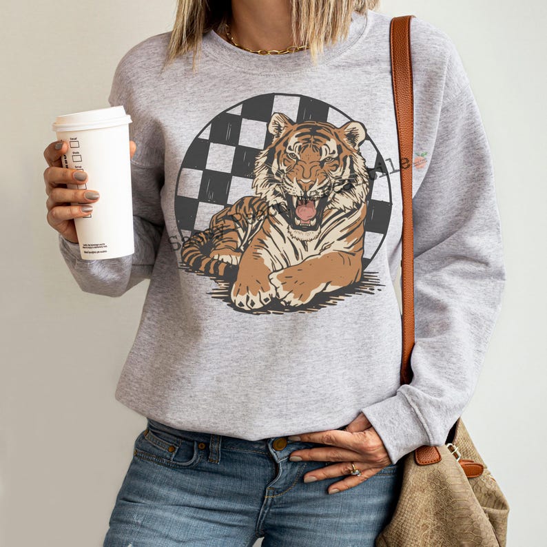 Puede incluir: Una sudadera gris claro con un gr&aacute;fico de tigre. El tigre es marr&oacute;n y beige, sobre un c&iacute;rculo a cuadros blanco y negro. La sudadera tiene mangas largas y cuello redondo.