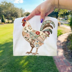 Könnte beinhalten: Weißes Sweatshirt mit einem farbenfrohen Hahn-Applikation. Das Hahn-Design enthält florale Muster und den Text "Peach Mercantile". Der Hahn hat einen roten Kamm und Kehllappen mit braunen und floral gemusterten Federn.