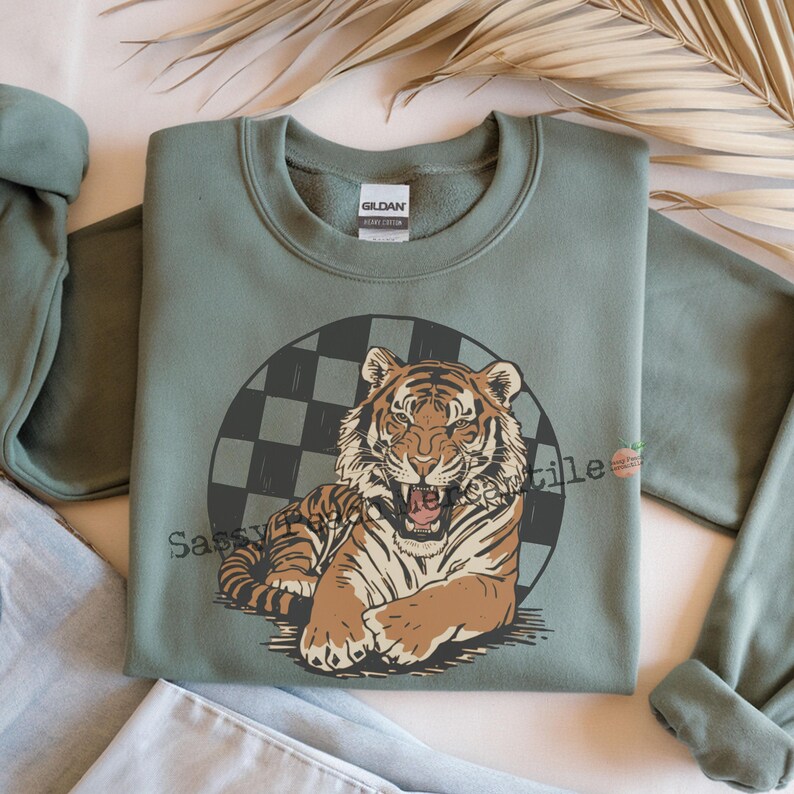 Puede incluir: Sudadera verde salvia con un gr&aacute;fico de tigre. El tigre, en tonos marr&oacute;n y beige, ruge, sobre un c&iacute;rculo a cuadros. El texto "Sassy" est&aacute; impreso en la parte inferior izquierda del gr&aacute;fico. La sudadera es de material suave.