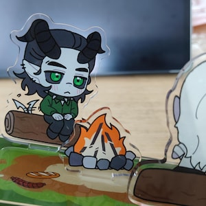 May include: A clear acrylic figurine depicts a cartoon character with horns, green eyes, and a green shirt, sitting on a log near a campfire. The base features a grassy area with food items. The overall theme is camping.