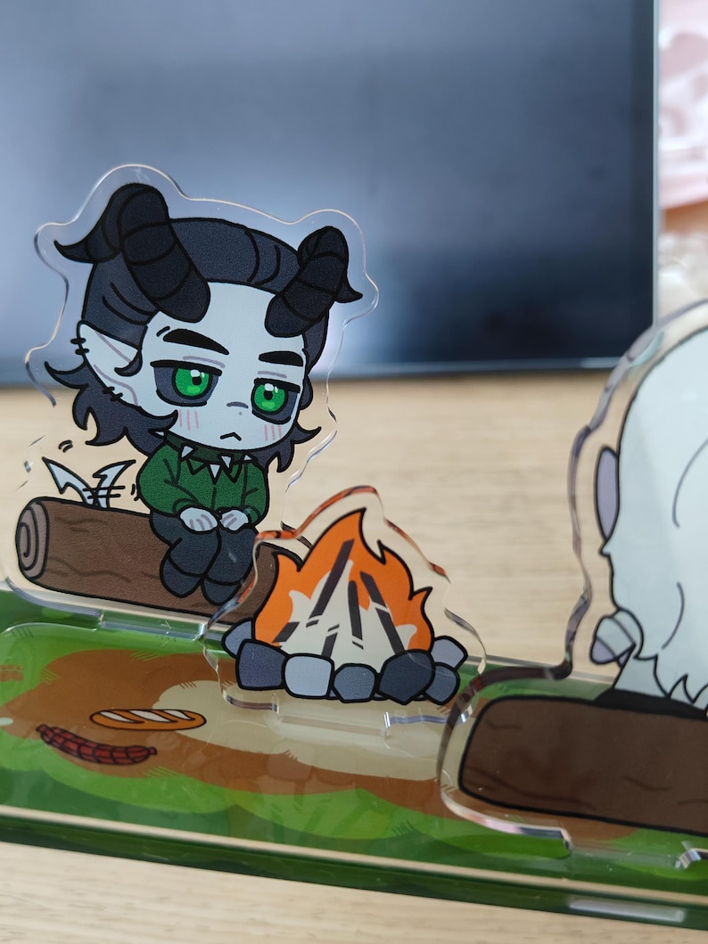 May include: A clear acrylic figurine depicts a cartoon character with horns, green eyes, and a green shirt, sitting on a log near a campfire. The base features a grassy area with food items. The overall theme is camping.