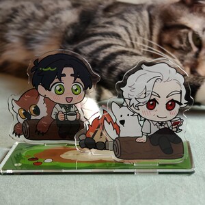 May include: Acrylic stand featuring two anime-style characters. One character is with an owl and a cup, the other with a white dog and a glass of wine. The stand has a green base with a painted landscape and a cat in the background.