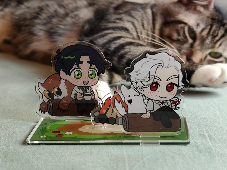 May include: Acrylic stand featuring two anime-style characters. One character is with an owl and a cup, the other with a white dog and a glass of wine. The stand has a green base with a painted landscape and a cat in the background.