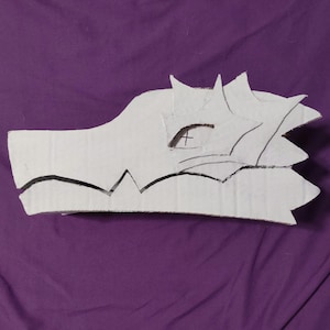 May include: A white cardboard mask shaped like a dragon head in profile. The mask features layered, jagged cutouts forming the eye area and a distinct, sharp jawline. A small black cross is drawn within the eye opening on the white surface.