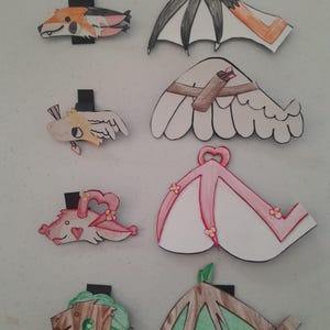 May include: A collection of colorful, handmade paper cutouts of fantasy creatures and wings. The designs include a fox, dragon, and bat wings, with details in orange, pink, black, and green. Each cutout is mounted on a black base.