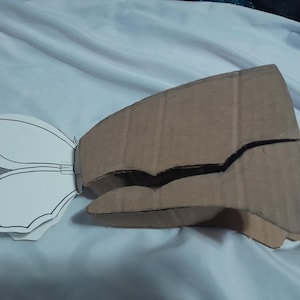 Puede incluir: Un proyecto de manualidades con una estructura de cartón corrugado marrón con forma de aleta o ala grande, unida a un recorte de papel blanco con el contorno negro de una forma de ala de mariposa. El cartón tiene costuras y capas visibles.