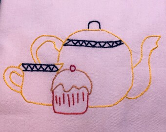 May include: Embroidered design of a yellow teapot, a yellow teacup, and a red cupcake with white frosting on a pink fabric background.