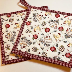 May include: Two quilted pot holders with a white background featuring a repeating pattern of chickens, red apples, and checkered pineapples. The pot holders have a red and white checkered border and a loop for hanging.
