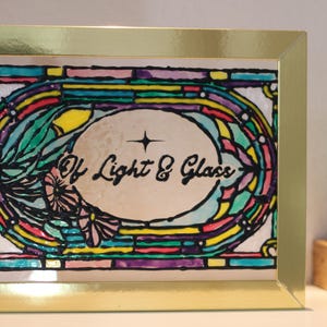 Puede incluir: Una obra de arte en vitral enmarcada con un marco dorado. La obra presenta un diseño colorido con el texto "Of Light & Glass" en el centro. El diseño incluye elementos florales y geométricos.