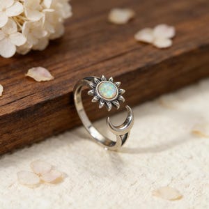 Puede incluir: Un anillo abierto de plata con un motivo de sol que tiene una piedra de ópalo iridiscente en el centro en un extremo y un motivo de media luna en el otro. El metal tiene un acabado oxidado para resaltar los rayos del sol y la curva de la luna.