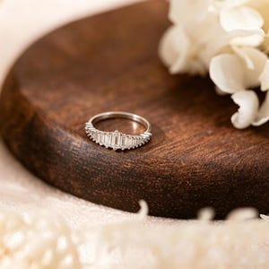 May include: A silver ring with a row of rectangular-cut stones in the center and smaller stones on either side. The ring is set on a dark wooden surface with white flowers in the background.