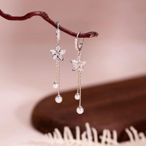 May include: Elegant silver earrings with a floral design. Each earring features a flower-shaped stud with dangling chains and three small, round, white pearls. The earrings are displayed on a dark brown branch against a soft pink background.