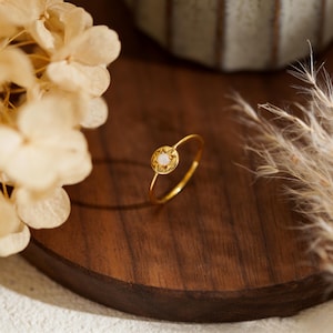 May include: A delicate gold ring with a small, round, off-white gemstone at the center. The ring's band is thin and smooth, and the gemstone is set within a starburst design. The ring is displayed on a dark wooden surface.