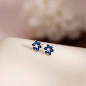 May include: A pair of flower-shaped stud earrings. Each earring features six dark blue gemstones surrounding a central white gemstone. The earrings are set on a light-colored surface with a soft, blurred background.