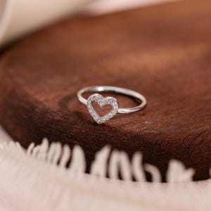 May include: A silver ring with a heart-shaped design. The heart is outlined with small, clear gemstones. The ring is set against a dark brown wooden surface, with a soft, white fabric in the background. The ring is a delicate piece of jewelry.