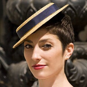 May include: A woman wearing a straw hat with a navy blue band. The hat is tilted at an angle and has a gold trim.