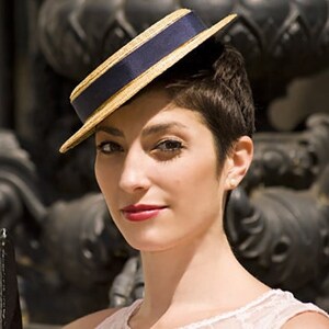May include: A woman wearing a straw hat with a blue ribbon and a white trim. The hat is tilted at an angle and the woman is looking at the camera.