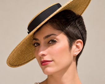 Puede incluir: Un sombrero de paja de ala ancha con copa plana y una cinta negra. El sombrero presenta una textura tejida en color beige natural y una silueta estructurada. La cinta negra rodea la base de la copa, creando un contraste con la paja.
