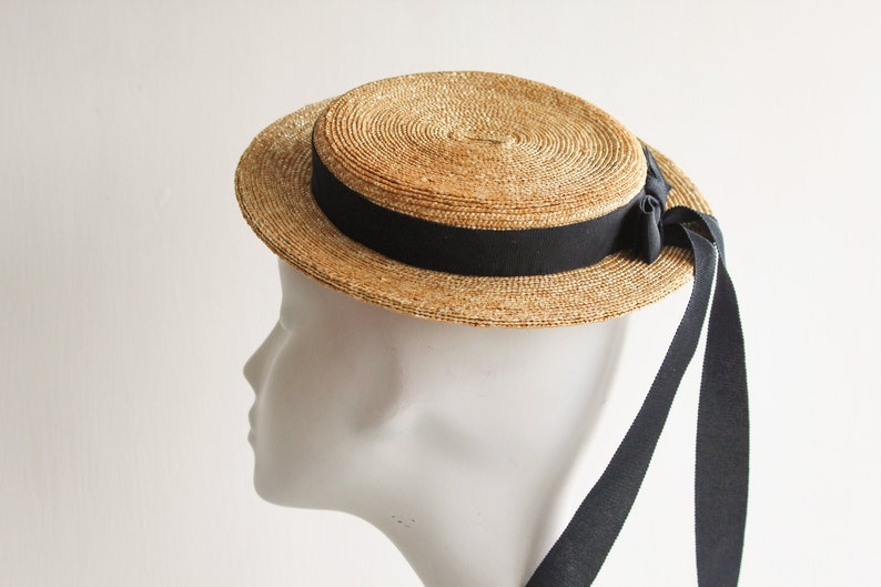 Puede incluir: Un sombrero de paja de tipo canotier con una cinta negra y largas cintas de lazo negras. El sombrero est&aacute; en una cabeza de maniqu&iacute; blanca.