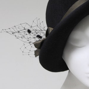 May include: A black felt hat featuring a dark metallic ribbon band tied in a bow on the side. A delicate black netting veil with small, dark, fuzzy circular accents is attached to the side of the hat, extending outward from the ribbon bow.
