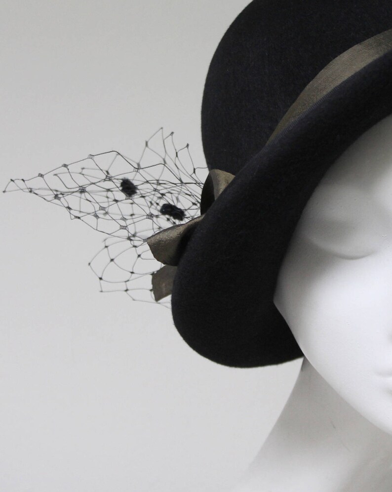 May include: A black felt hat featuring a dark metallic ribbon band tied in a bow on the side. A delicate black netting veil with small, dark, fuzzy circular accents is attached to the side of the hat, extending outward from the ribbon bow.