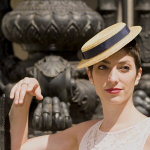 May include: A woman wearing a straw hat with a blue band and a white lace dress. She is standing in front of a black metal structure.
