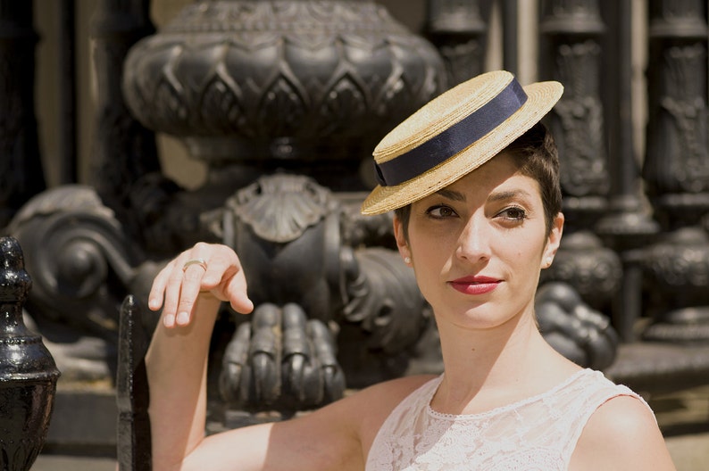May include: A woman wearing a straw hat with a blue band and a white lace dress. She is standing in front of a black metal structure.