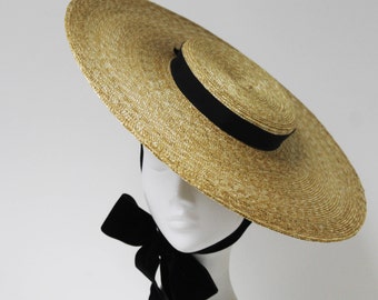 Puede incluir: Un sombrero de paja de ala ancha con una cinta negra alrededor de la corona. El sombrero está inclinado y tiene un lazo de terciopelo negro atado en la parte posterior.