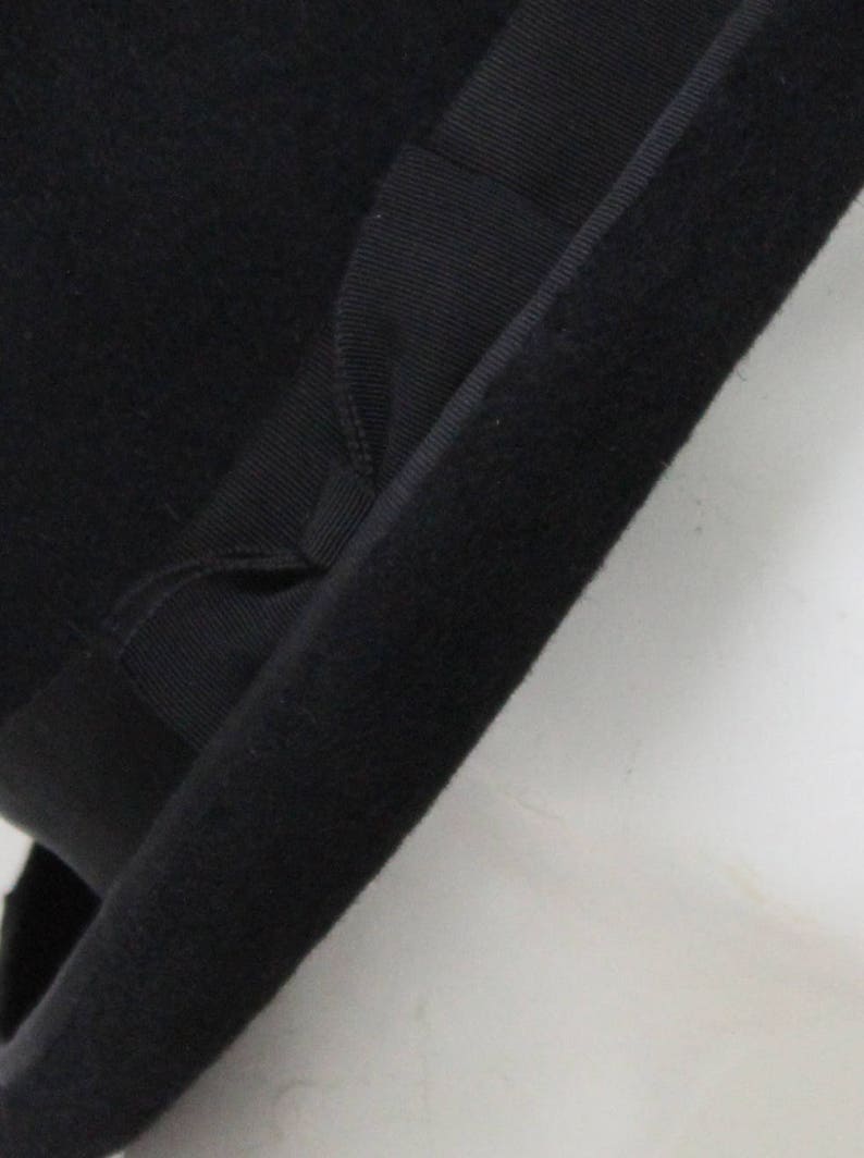 May include: A black felt hat featuring a wide, structured brim and a matching black grosgrain ribbon band wrapped around the base of the crown. The material has a soft, matte texture, and the ribbon includes a small, folded bow detail.