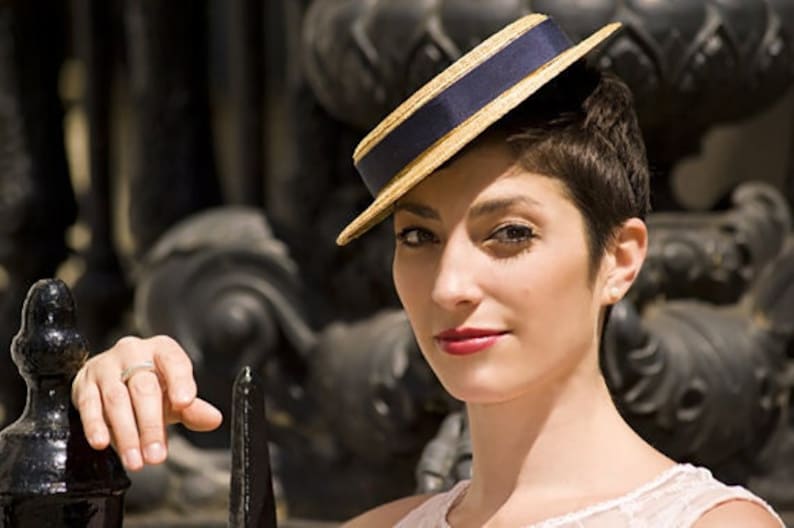 May include: A woman wearing a straw hat with a blue ribbon and a white trim. The hat is tilted at an angle and the woman is looking at the camera.