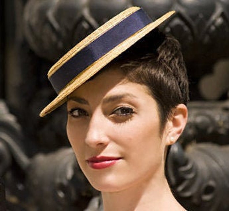 May include: A woman wearing a straw hat with a navy blue band. The hat is tilted at an angle and has a gold trim.