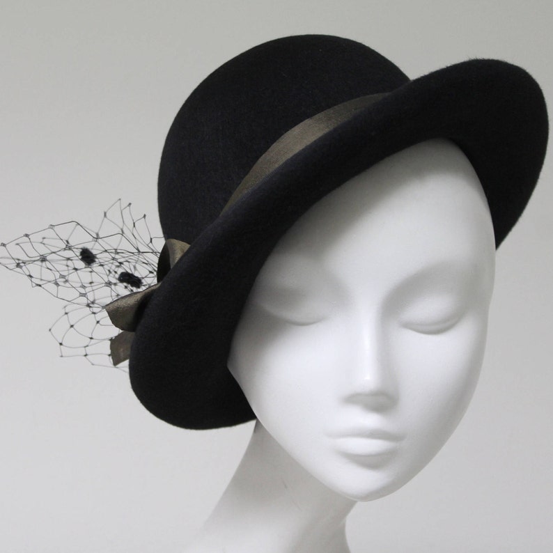 May include: A black felt hat with a rounded crown and a curved brim. The hat features a dark brown ribbon band tied in a bow on the side, accented with a black netting veil that extends outward from the bow, adding texture to the solid black design.