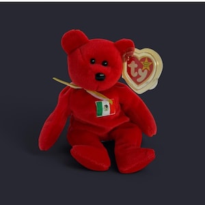 Pode incluir: Um urso de pelúcia vermelho com nariz e olhos pretos. O urso tem um pequeno patch da bandeira mexicana no peito e uma etiqueta em forma de coração Ty. O urso é um item de colecionador.