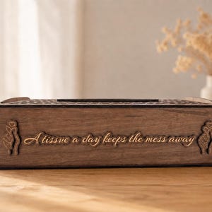 May include: A rectangular dark brown wooden tissue box cover featuring the laser-engraved text "A tissue a day keeps the mess away" on the front panel, flanked by two small engraved hand heart symbols on the left and right sides.
